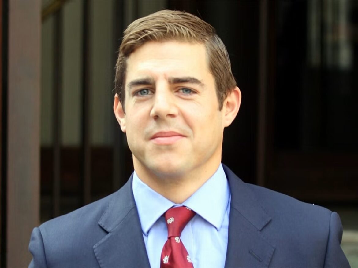 Portrait of a man in a navy suit and light blue shirt with a red patterned tie, standing outdoors and looking at the camera, smiling subtly.