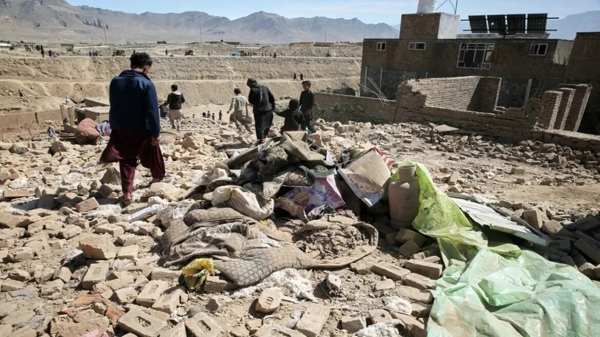 Group of people sifting through rubble in a dusty desert town, with a pile of mattresses, bags, and a green tarpaulin amid brick debris and distant mountains.