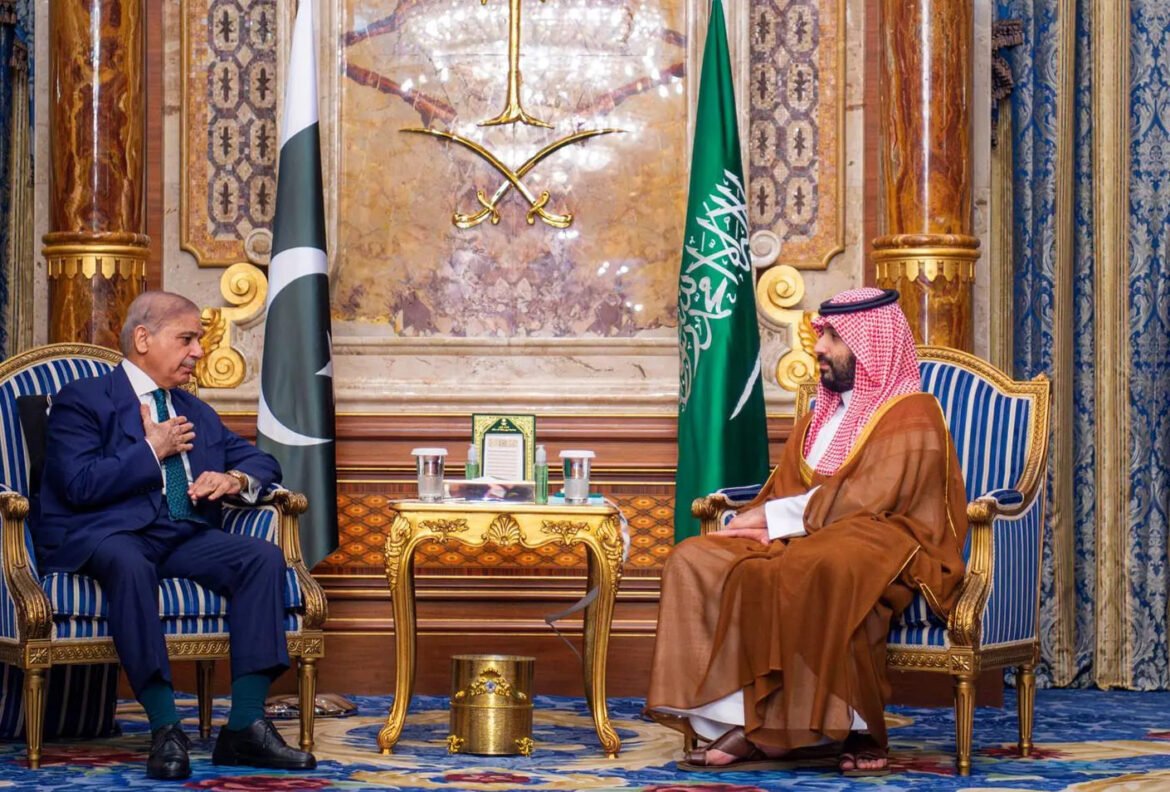Two men in formal attire sit in ornate gold-trimmed chairs across from each other in a gilded room with Pakistan and Saudi flags behind them, signaling a high-level meeting.