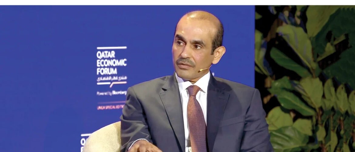 Male speaker in a dark suit and maroon tie on stage at the Qatar Economic Forum, blue backdrop.