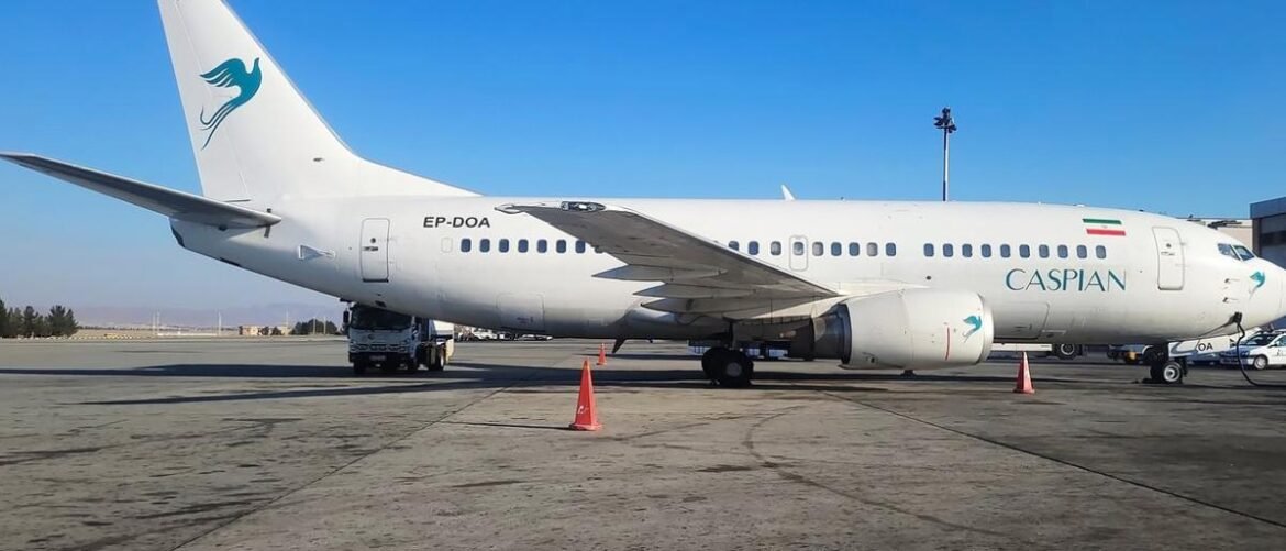 White Caspian Airlines jet on tarmac, tail logo of a blue bird, Iranian flag near rear, engines visible under wing.