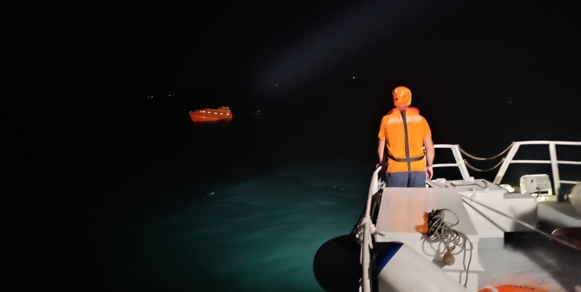 Back view of a person in an orange life jacket standing on the right side of a boat at night, looking toward a distant orange lifeboat on dark water.