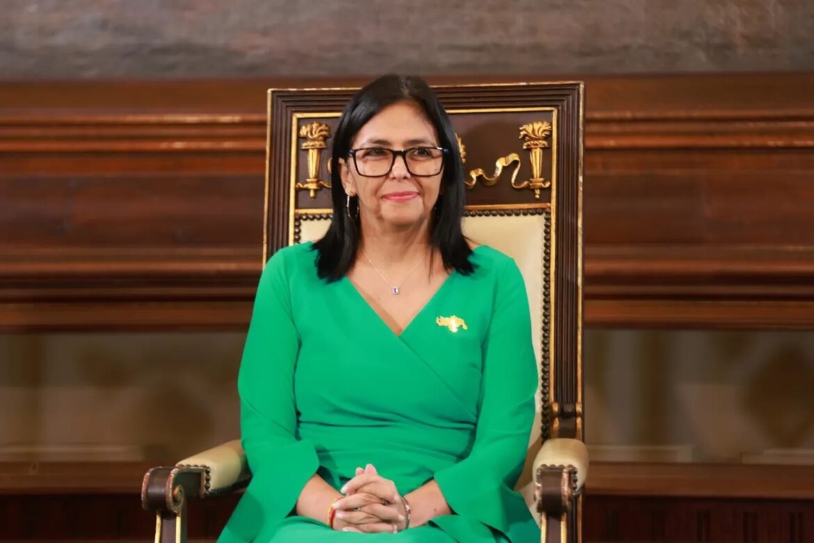 Woman with black hair and glasses seated in an ornate wooden chair wearing a green dress in a formal room setting.