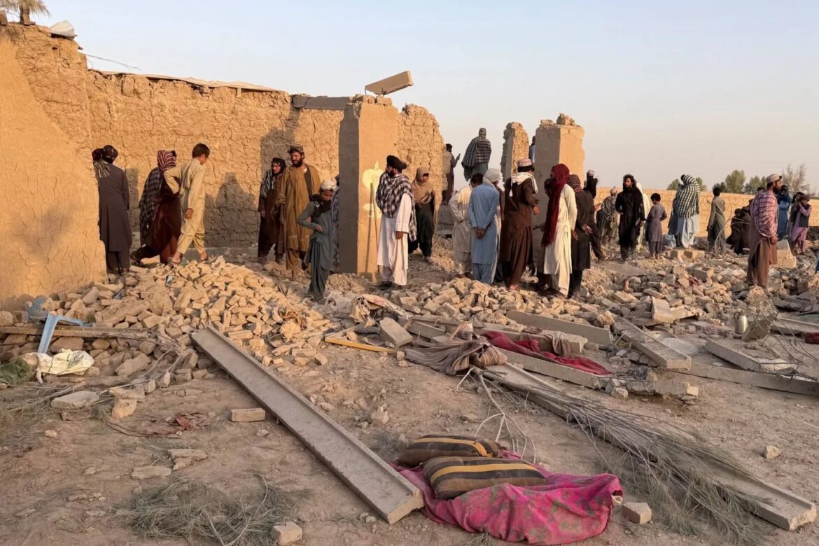 People stand among rubble and ruined mud-brick buildings after a collapse, surveying debris and makeshift tents.
