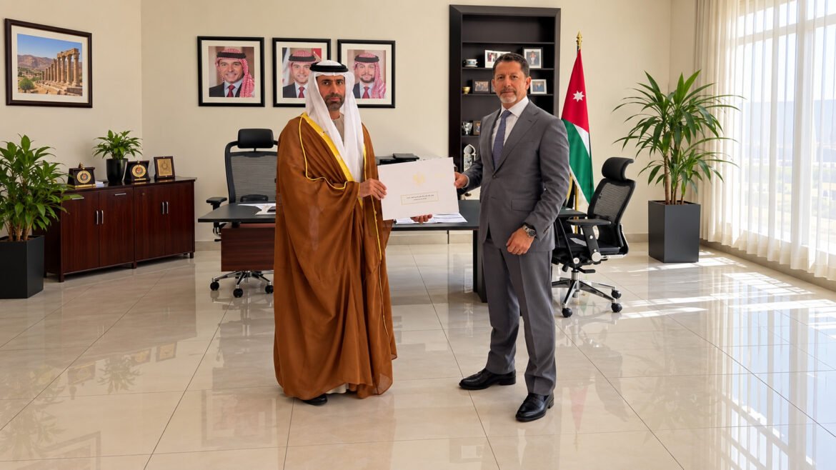 Two men in an office exchange a certificate; one wears a brown thobe and white headscarf, the other a gray suit, with the Jordan flag in the background.