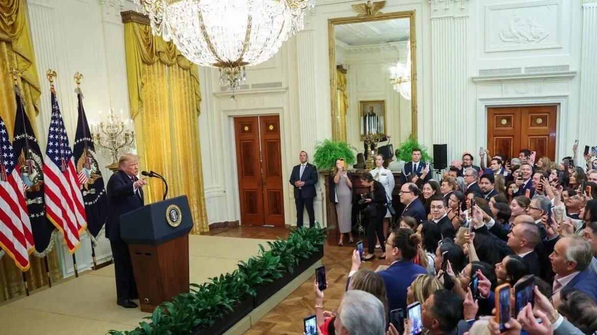 President at a podium giving a speech in a formal hall with American flags and a large chandelier, addressing a crowd taking photos with phones.