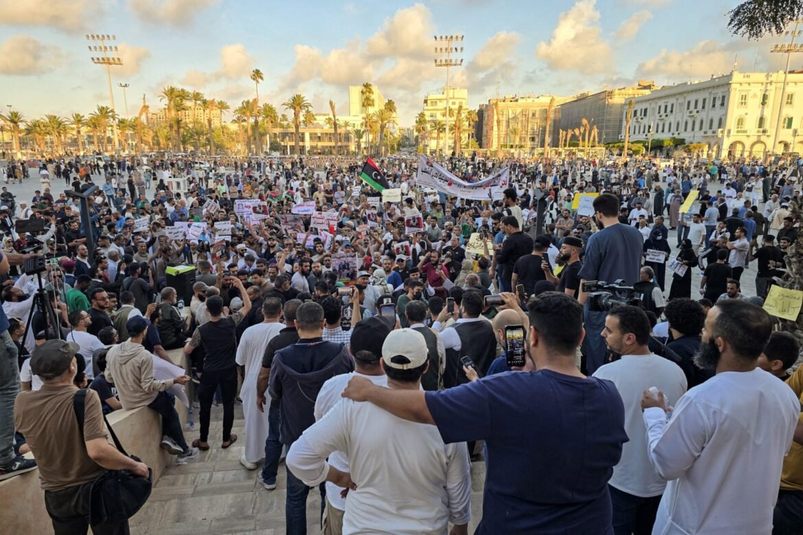 Massive crowd of protesters in a sunny square, holding banners and a Libyan flag, with palm trees and historic buildings behind.