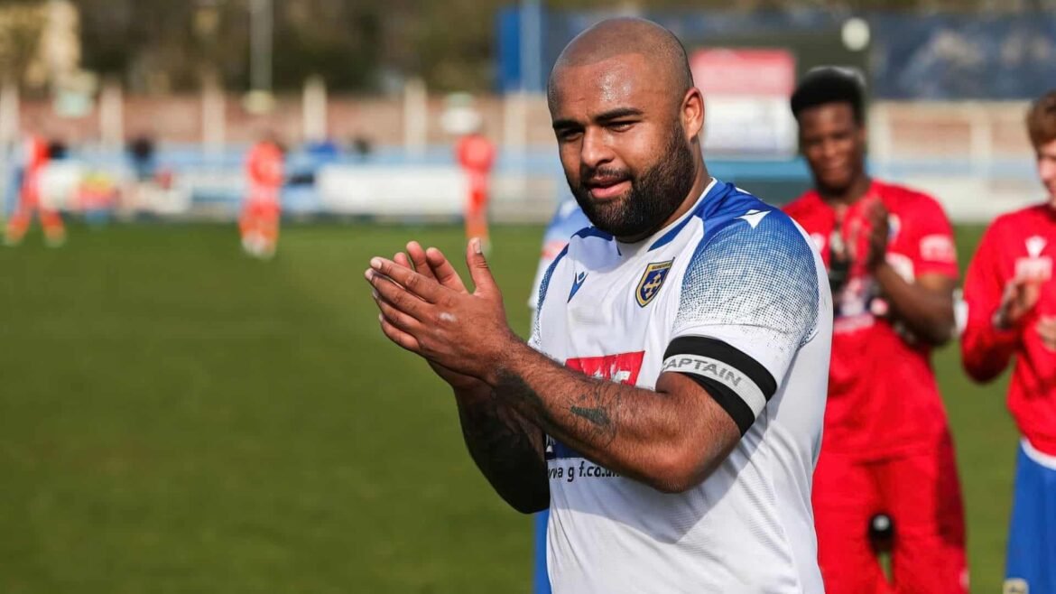 Soccer player in white-blue jersey with captain armband clapping on a sunny field; teammates in red in the blurred background