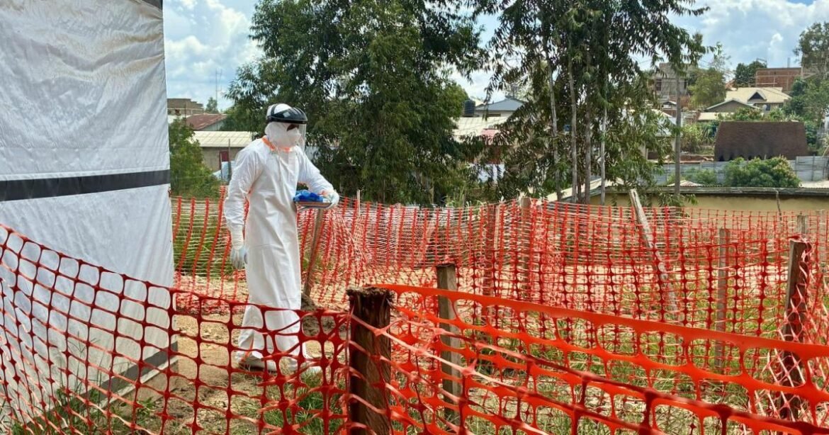Worker in full protective suit and face shield walking along an orange safety fence at a construction or quarantine site.