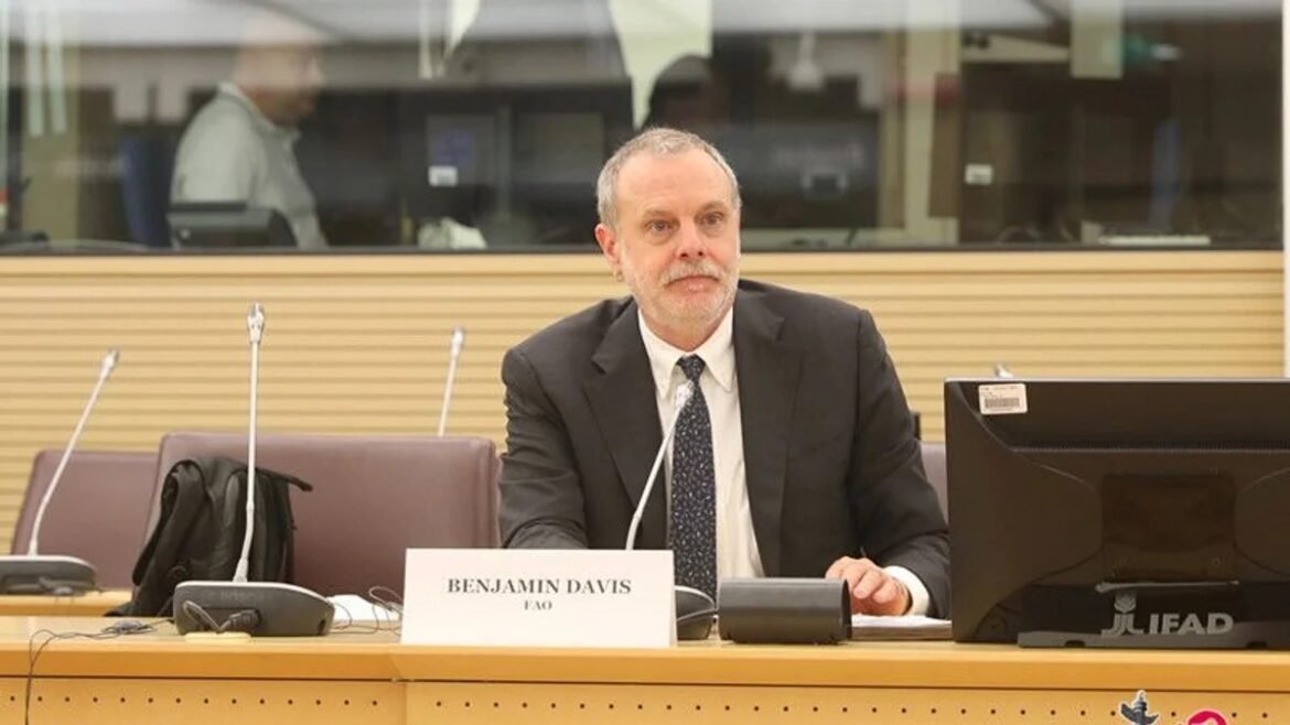 Man in a dark suit and tie sits at a conference desk with a nameplate reading 'Benjamin Davis' and a computer monitor to the right.
