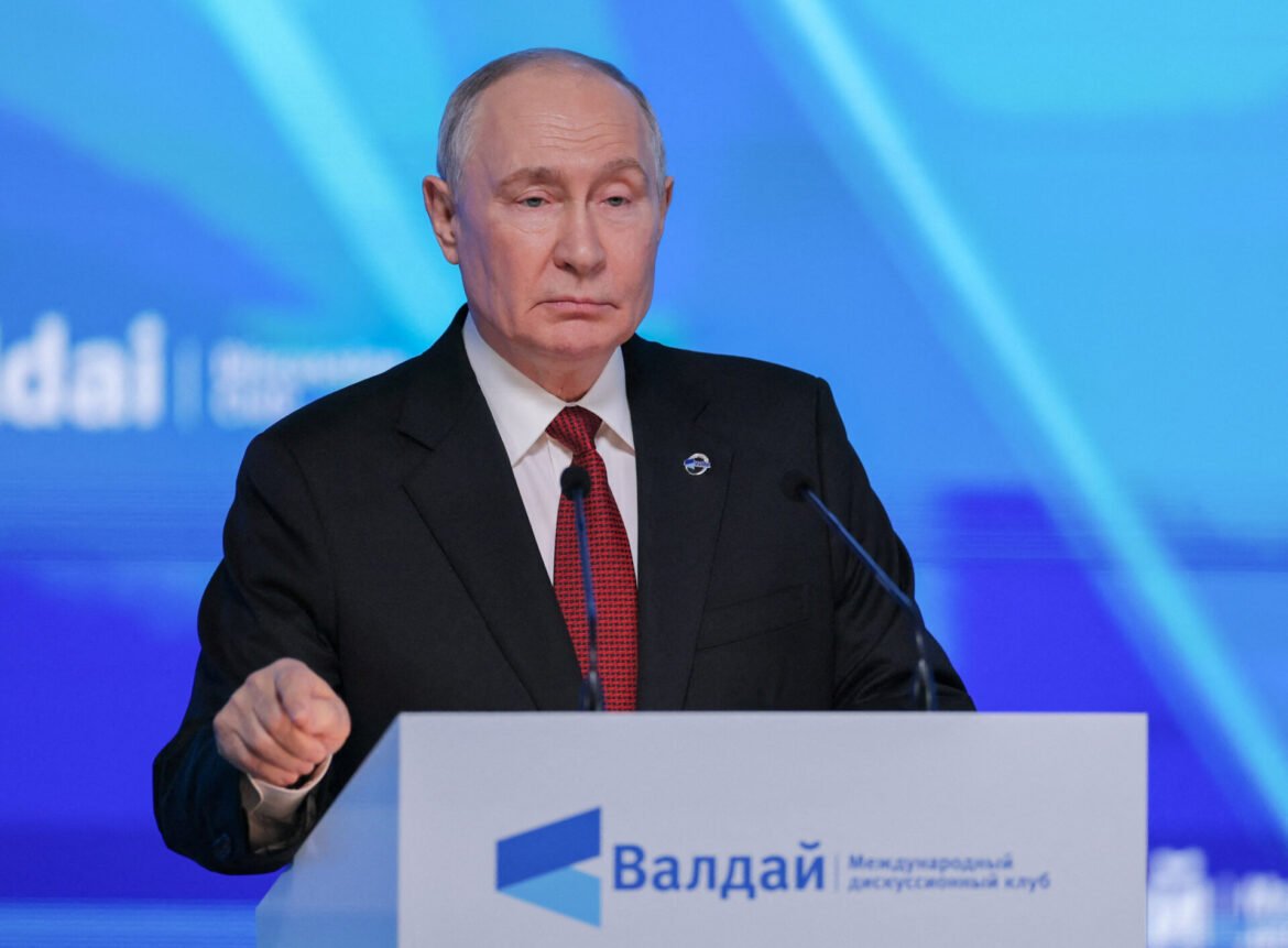 Male speaker in a black suit and red tie standing at a lectern with a blue backdrop behind him. The podium bears a logo and Cyrillic text.