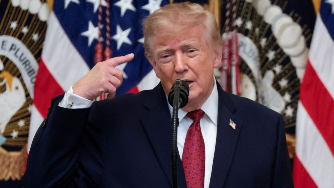 Donald Trump speaking at a podium with a microphone, gesturing toward his temple with flags in the background.