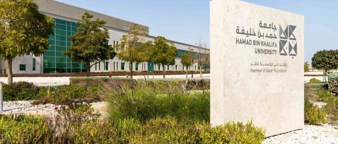 Stone sign at Hamad Bin Khalifa University with Arabic text; campus building with green glass windows in background.